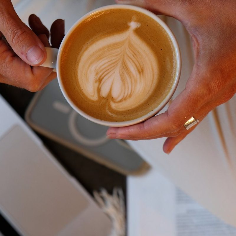 Hände halten einen Cappuccino mit Latte Art über Laptop und Buch
